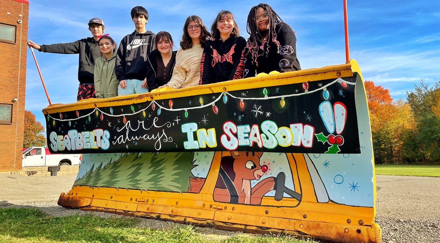 Yellow snow plow painted for the Holidays with a tree and deer with lights. Students in background.