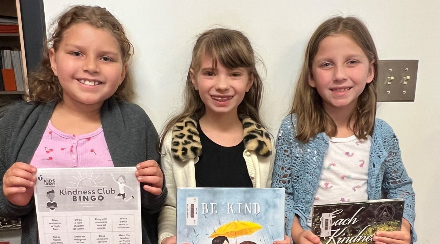 White wall background.  3 students holding up books about Kindness.