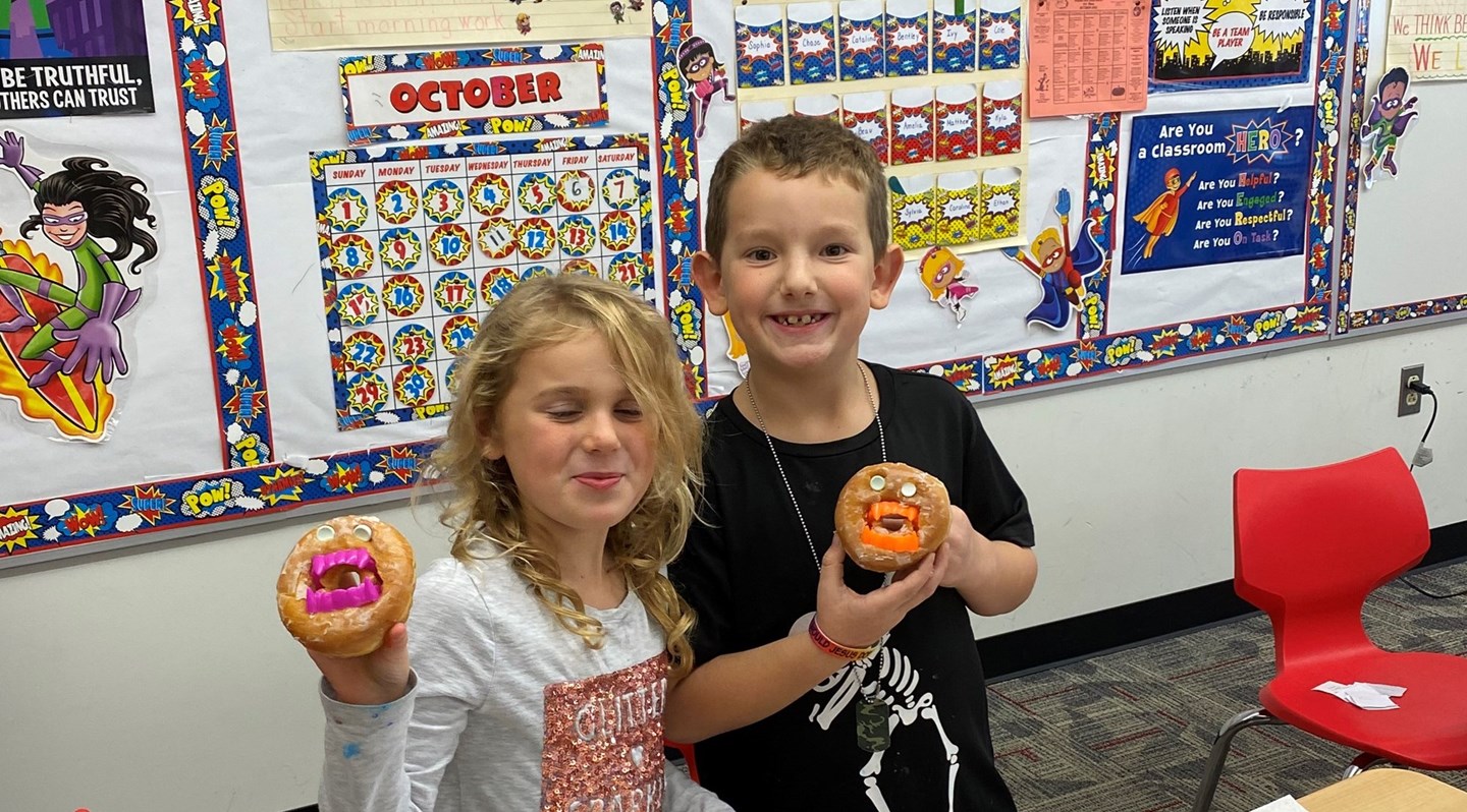 Two Students holding up  donuts with vampire teeth on the middle and eyes.