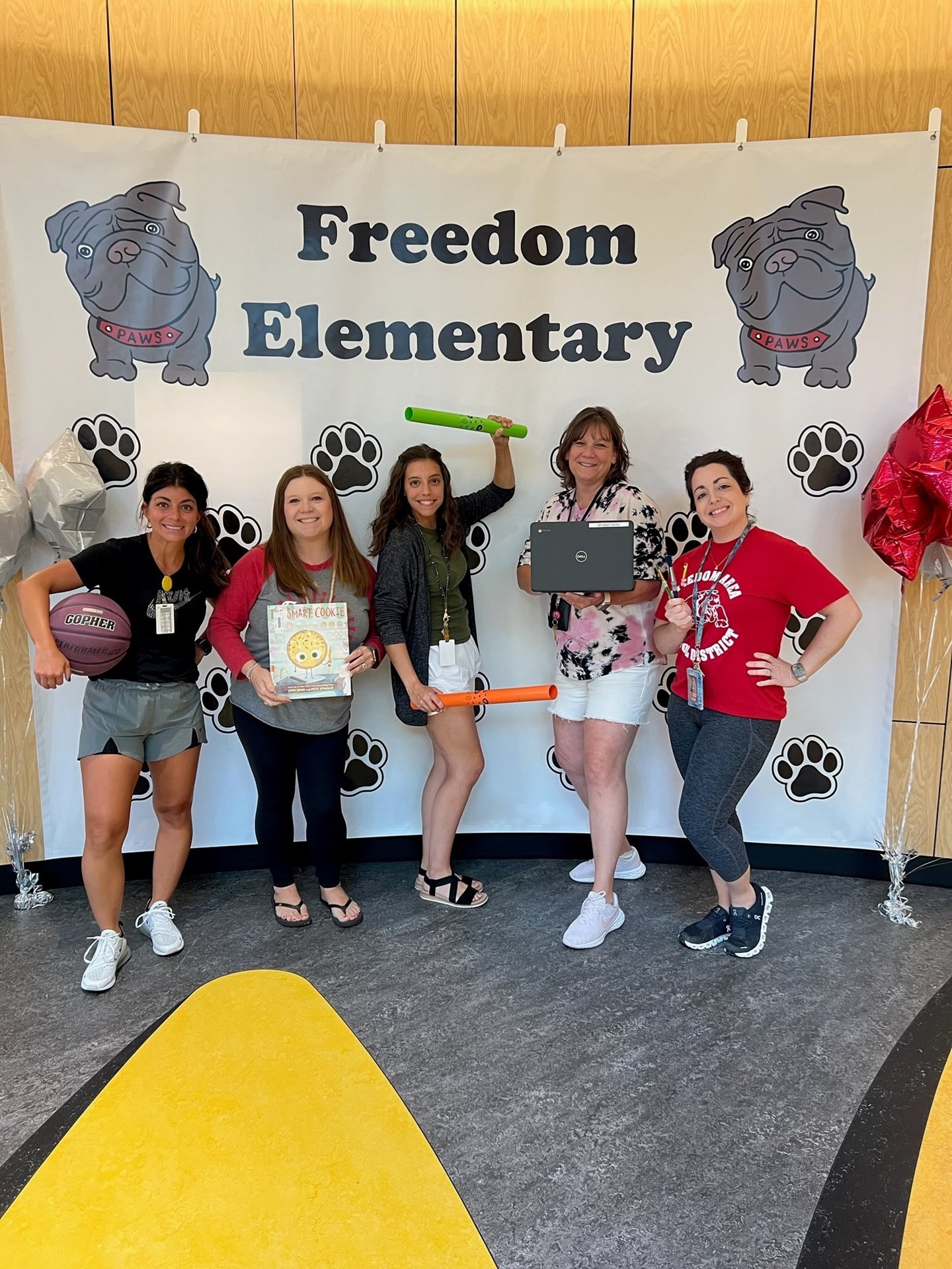 Two gray bulldogs on the top corners. Black pawprints in the back ground.  Picture of teachers. first on is holding a basketball, second is holding a white book, third is holding a green and orange stick, forth is holding a computer and last is holding a paint brush.