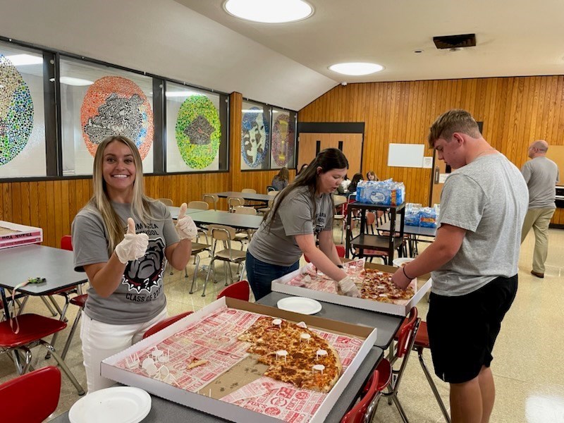 Cafeteria with gray tables and Red Chairs.  Pizza in pizza boxes.