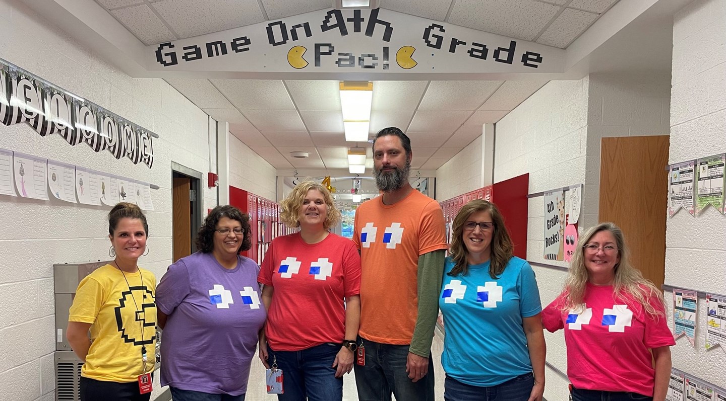 6 teachers in the hallway of a school with white walls. Each teacher as on different colors of pacman shirts