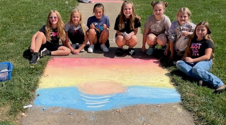 Red brick school in background.  Green grass.  Gray sidewalk with kids and chalk on it.